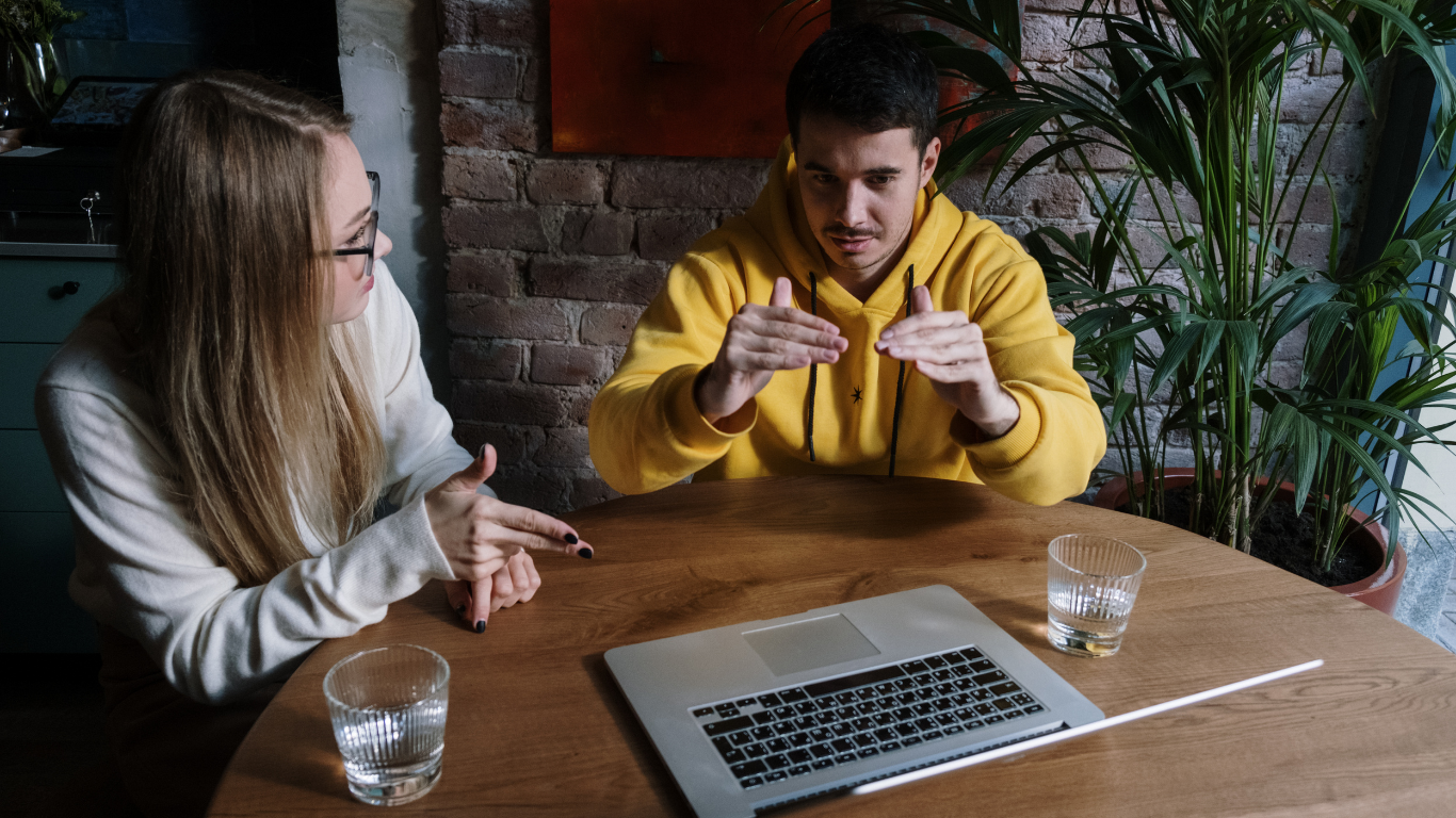 Zwei Menschen sitzen an einem Tisch, vor ihnen steht ein Laptop und sie unterhalten sich miteinander